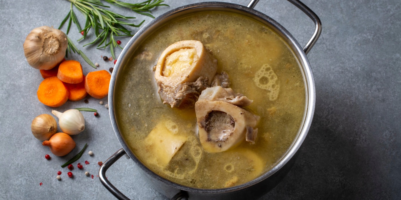 A steel pot of golden liquid, bones and other ingredients on a counter with some garlic, onion, carrots and rosemary. 