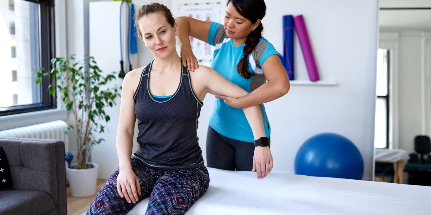 A woman sits on a therapy table, as a physiotherapy practitioner works on her shoulder.  A blue yoga ball is blurred in the background along with an indoor plant, yoga mats and tension cords. 