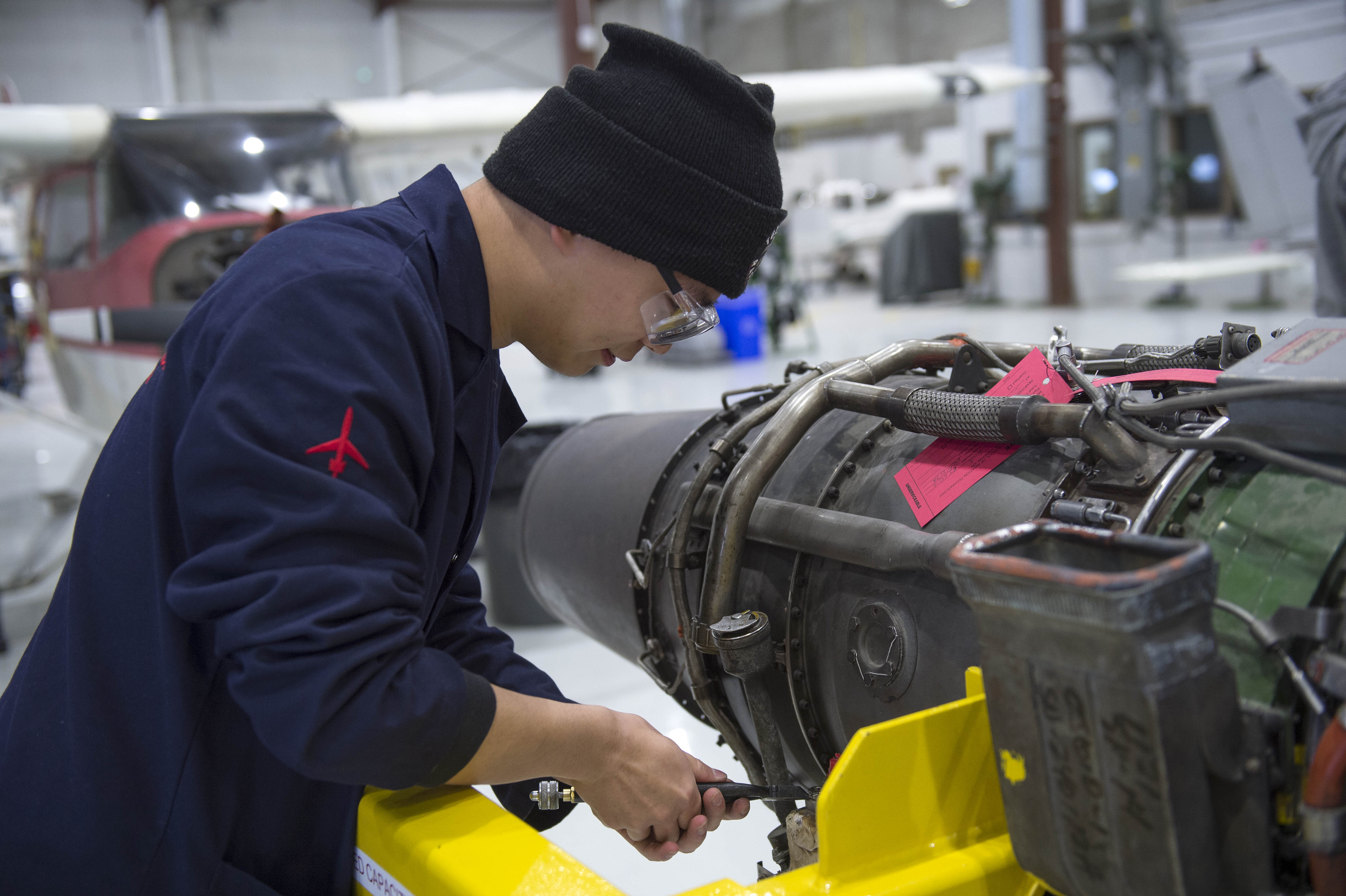 At Fanshawe College's Aviation Centre, a person wearing safety glasses and a navy uniform with a red airplane logo on the sleeve works on an aircraft engine mounted on a yellow stand.