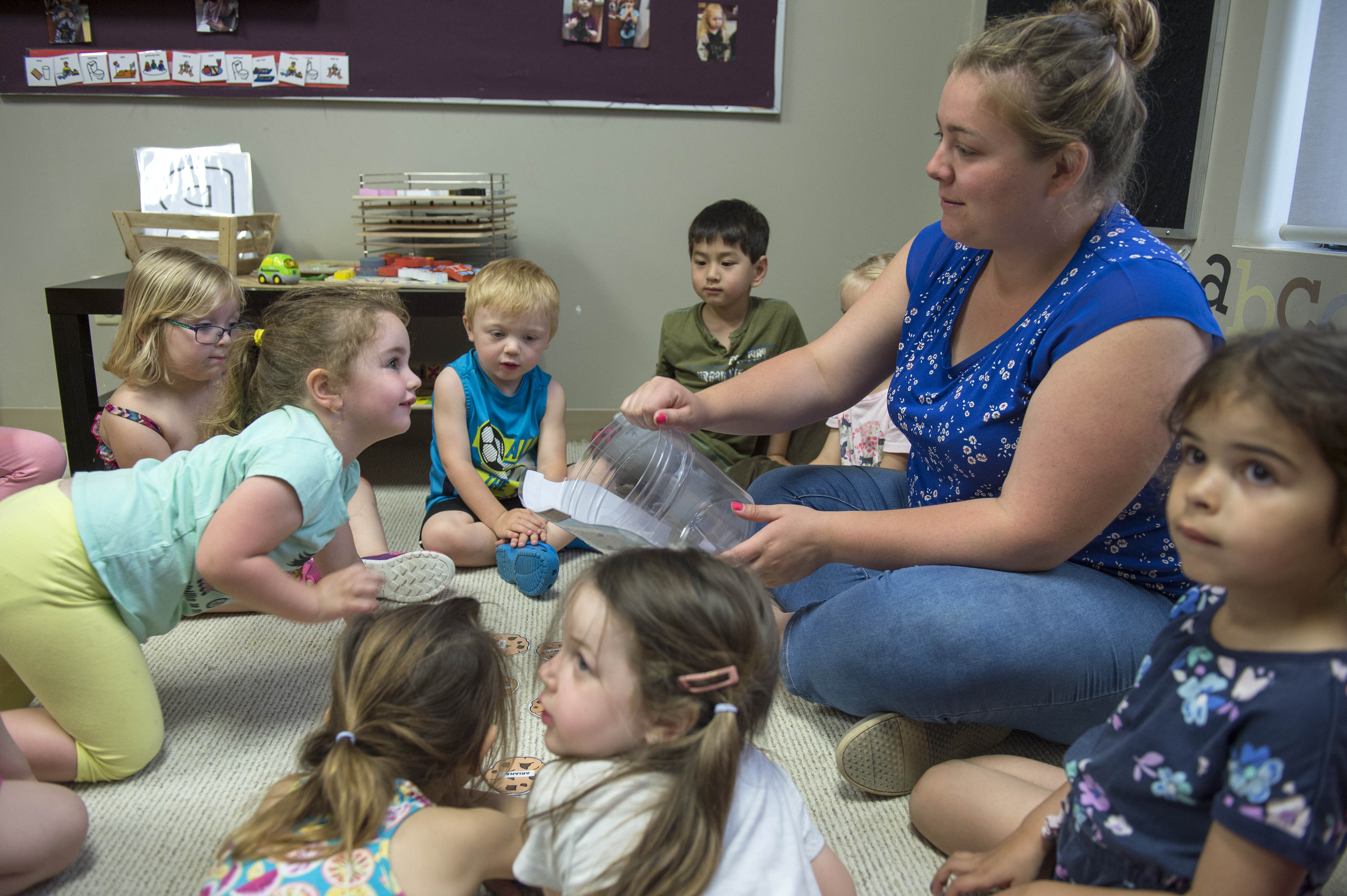 In a classroom at the Simcoe/Norfolk Regional Campus, an educator sits cross-legged on the floor, holding a clear plastic container, surrounded by children who are also sitting on the floor. The children are looking at the container and each other. Behind them is a low table with toys and learning materials, and a bulletin board with pictures and visual aids. The letters "abc" are visible on the wall. This is likely an activity for the Early Childhood Education program.