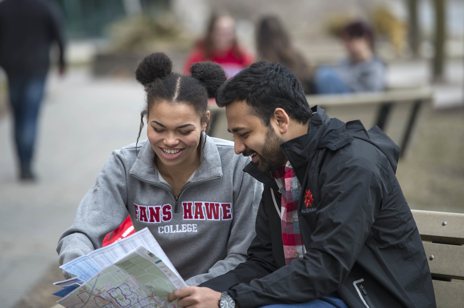 Two people wearing Fanshawe College branded clothing look at a map while sitting on a bench at Fanshawe College's London Campus.