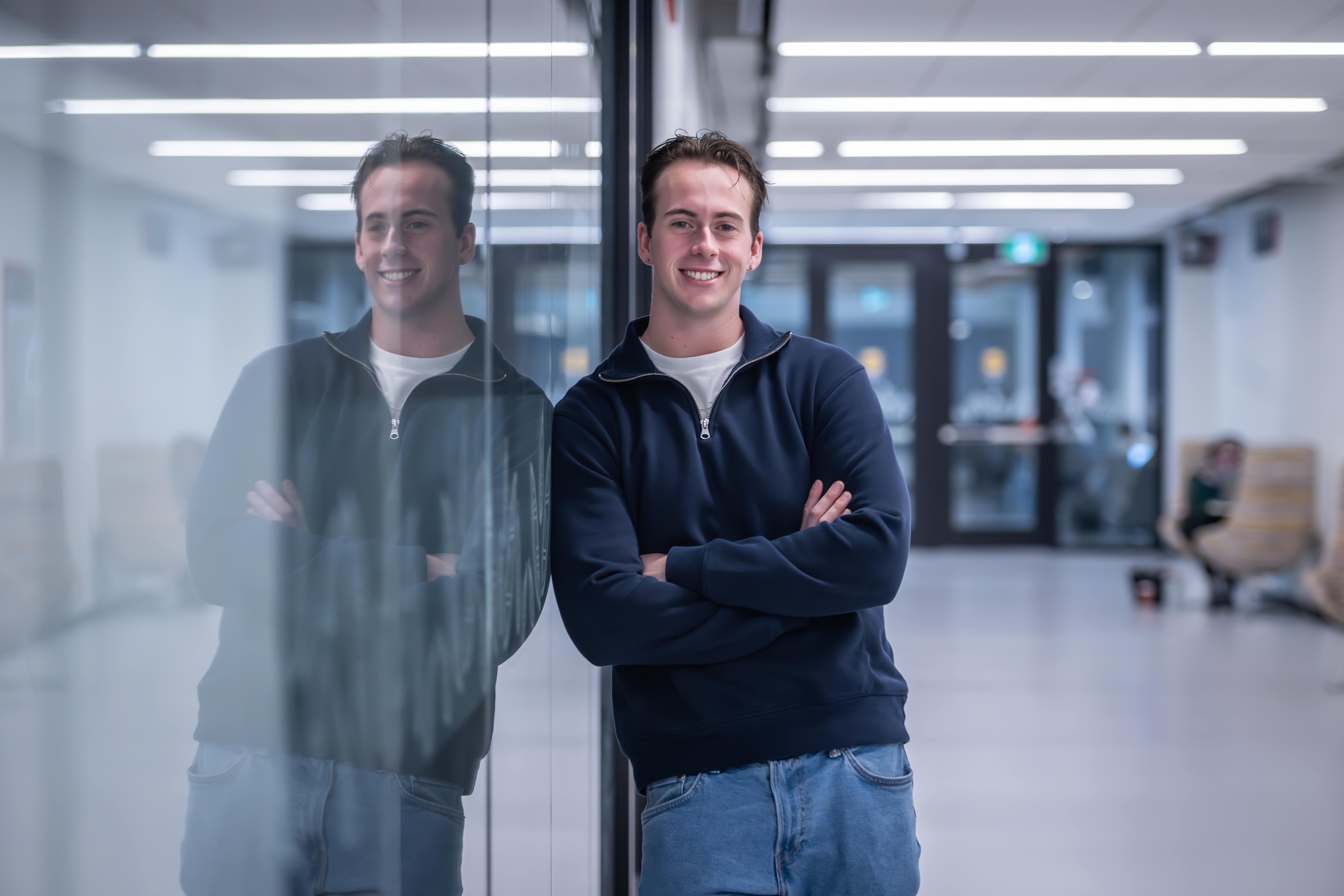 A person in a navy sweater and jeans stands with arms crossed, leaning against a glass wall that reflects their image.