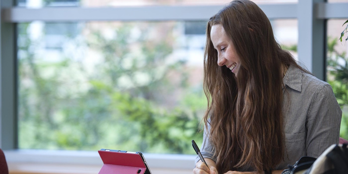 Student writing at table while looking at tablet