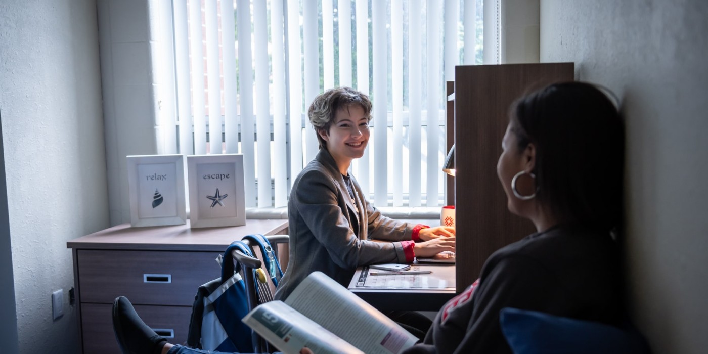 Woman doing homework smiling at roommate who is sitting on bed
