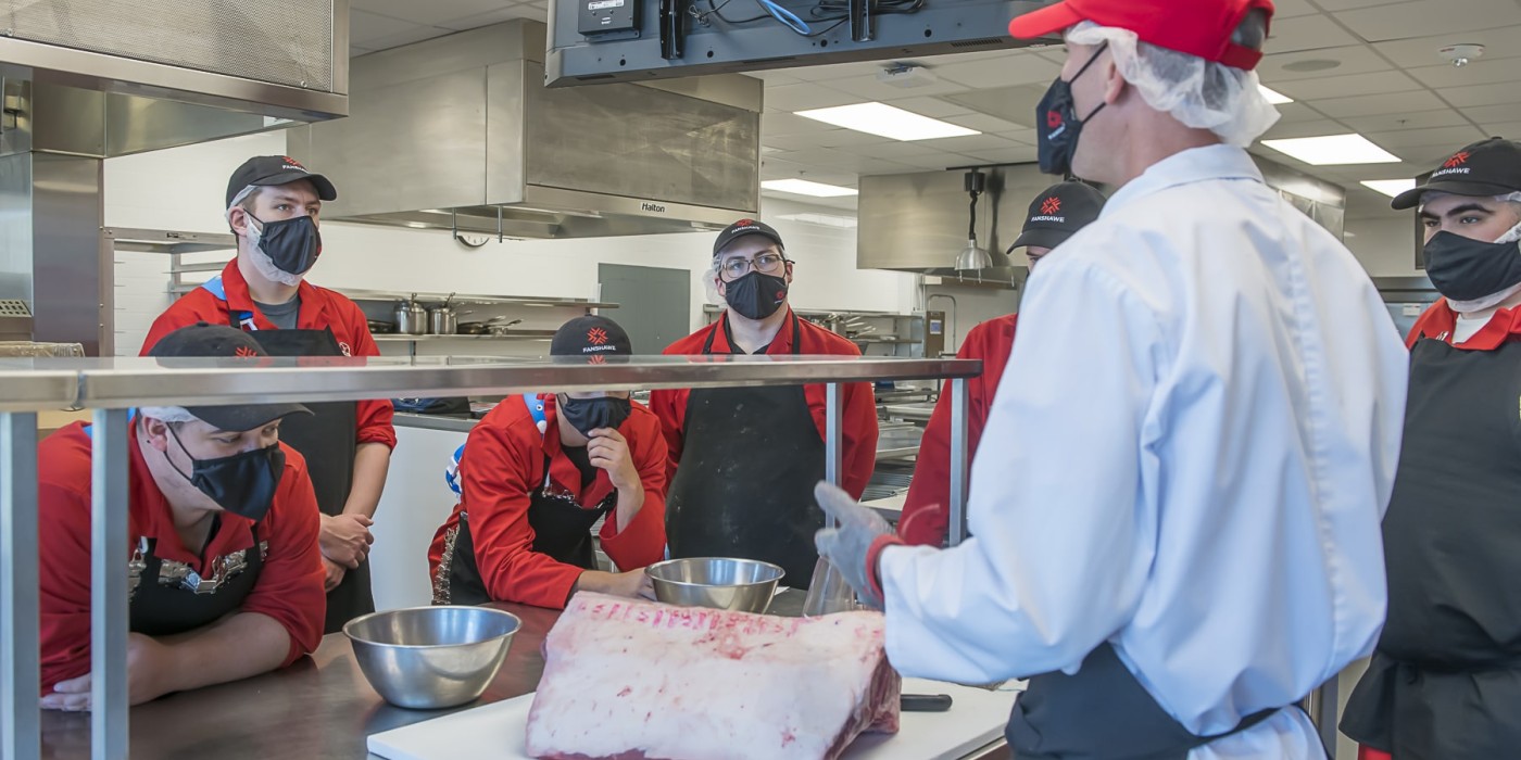 man stands in front of butcher counter talking to butchery students