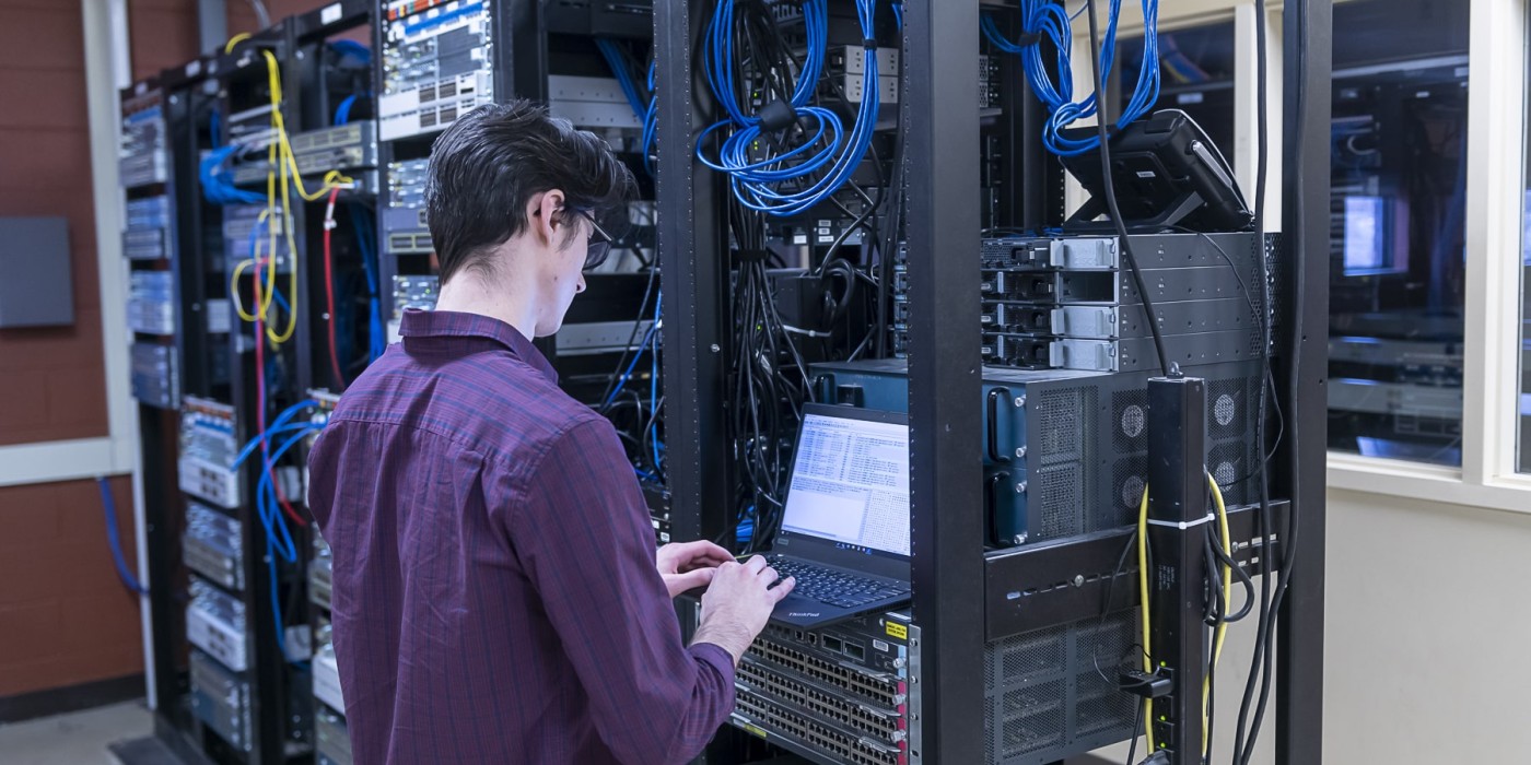 man stands in front of servers while typing on a laptop