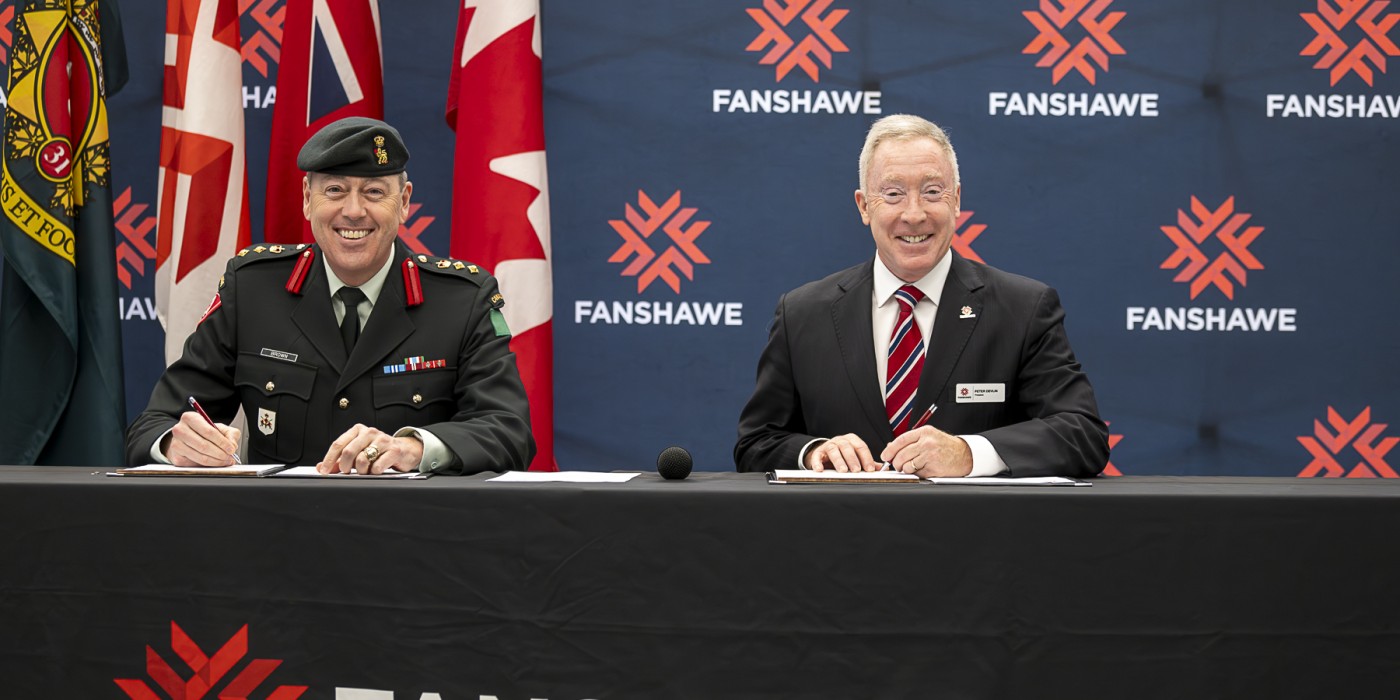 Colonel Chris Brown and President Peter Devlin seated at a table in front of Fanshawe backdrop to sign an MOU agreement.