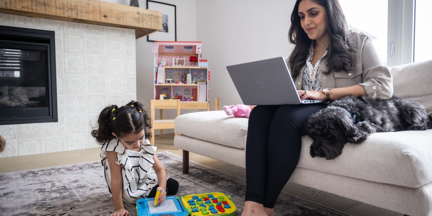mother sits on couch studying on laptop while child plays nearby