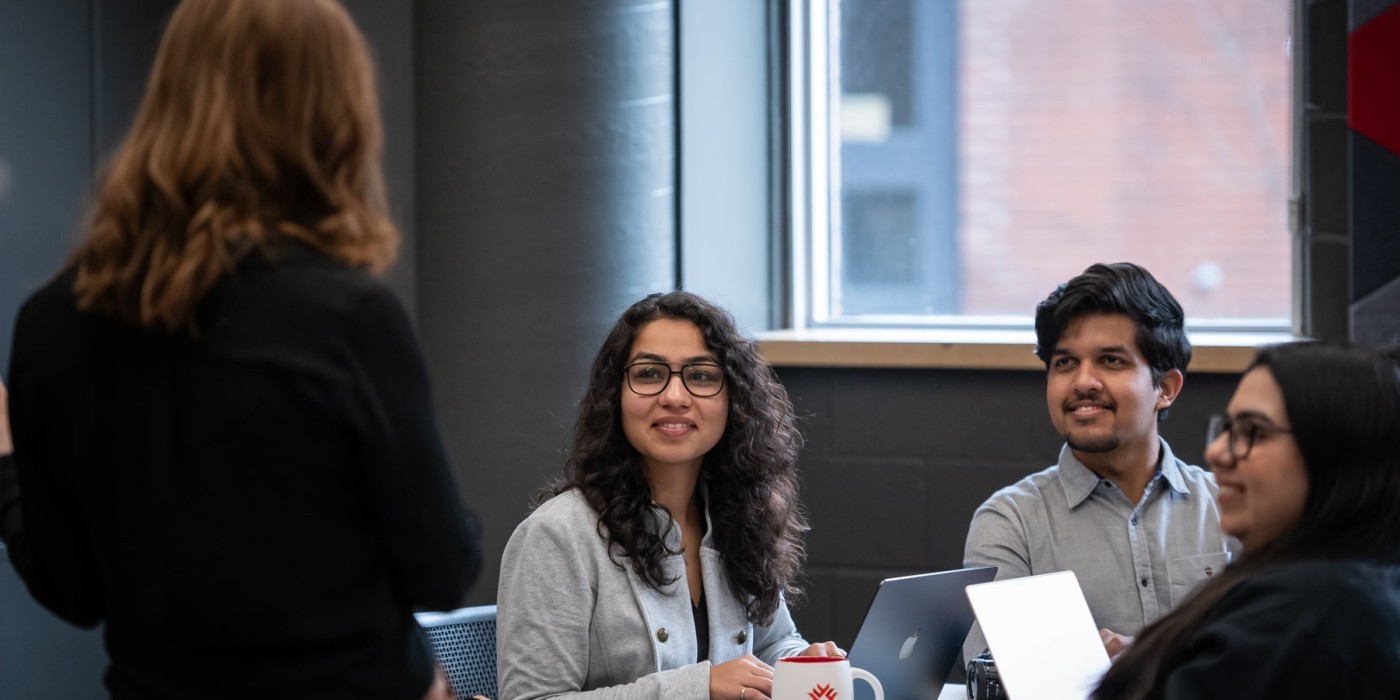 students sit at table with laptops, listening to presenter