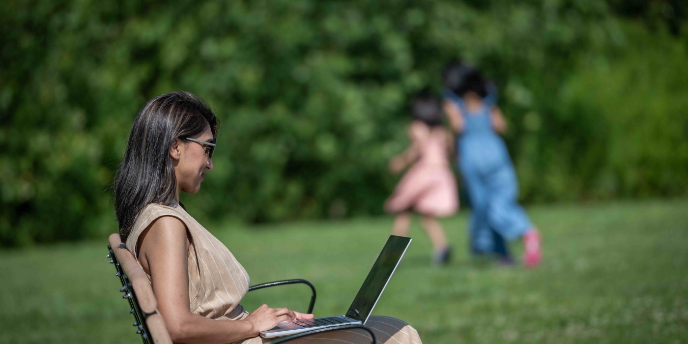 student sits outside on laptop while children play in background