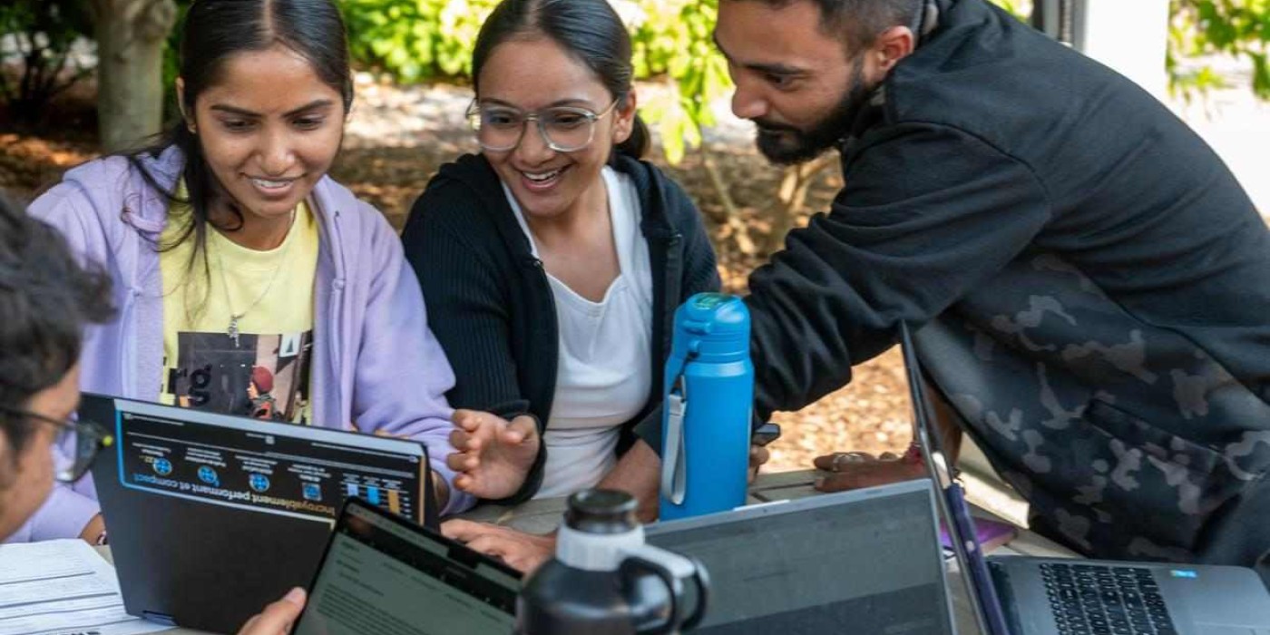 students sit around an outdoor table, pointing at laptop