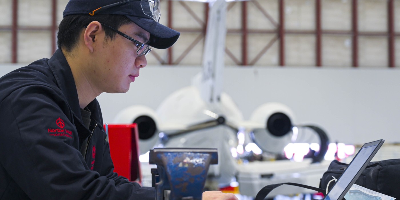 Aviation student sitting at a laptop with a plane in the background.