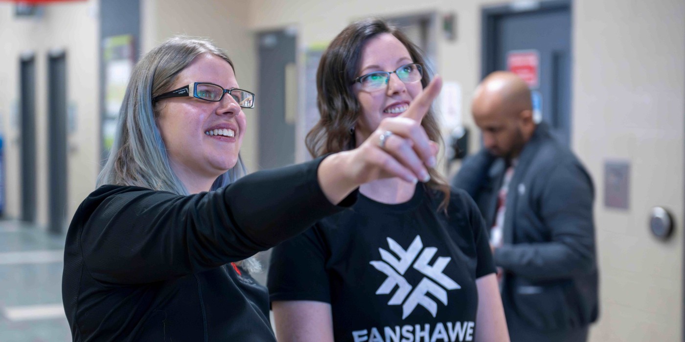 two people stand together at Fanshawe, pointing to a tv above them