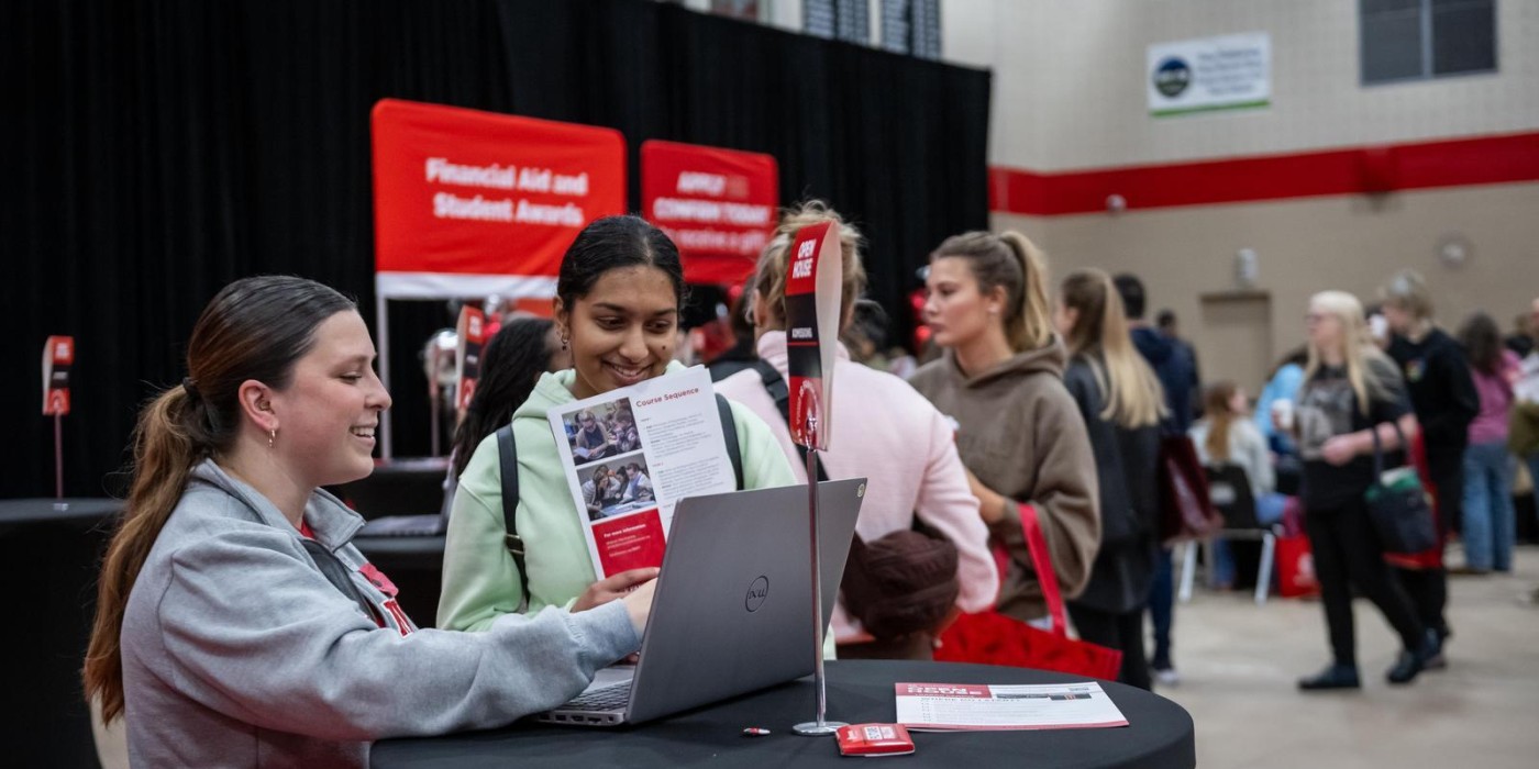 At the Fanshawe College London Campus Open House, two people are looking at a laptop and a brochure at a table with a black tablecloth, with a red banner in the background that reads "Financial Aid and Student Awards," and other people are smilling around in the background.