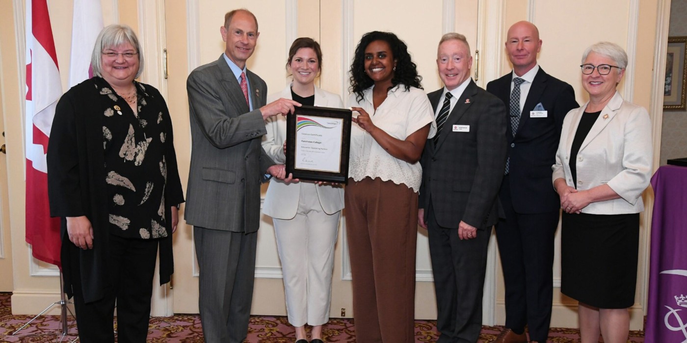 A group of seven people, including His Royal Highness Prince Edward The Duke of Edinburgh, are lined up smiling at the camera next to a Canadian flag.