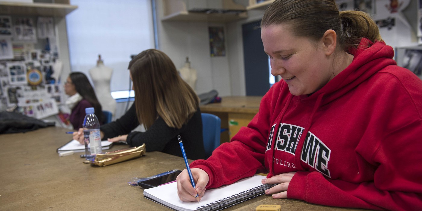 At the London Campus of Fanshawe College, three people sit at a long table, writing in notebooks with pencils, in a Design Foundations program classroom.