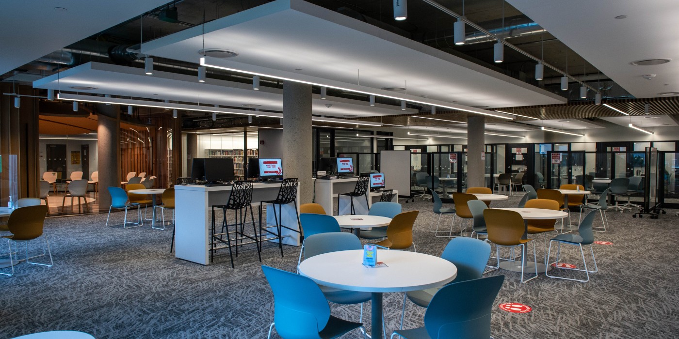 A study area with round tables and chairs is illuminated by overhead lighting. Computer workstations with high stools are visible in the background.