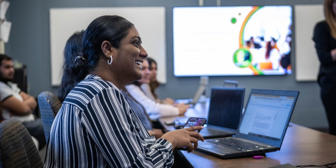 At Fanshawe College's London Campus, a business class is in session, with a person in a striped shirt smiling while holding a phone, surrounded by laptops on a table, and a presentation