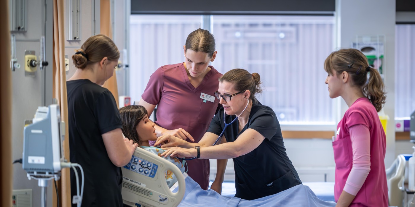 Four individuals in medical scrubs surround a patient simulator in a hospital room, one using a stethoscope.