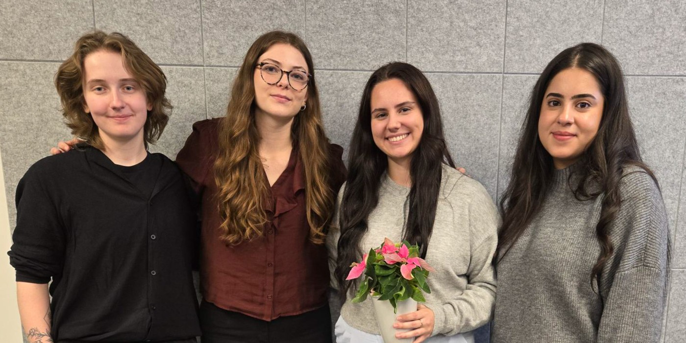 Four students stand in a line smiling at the camera. One student holds a flower pot.