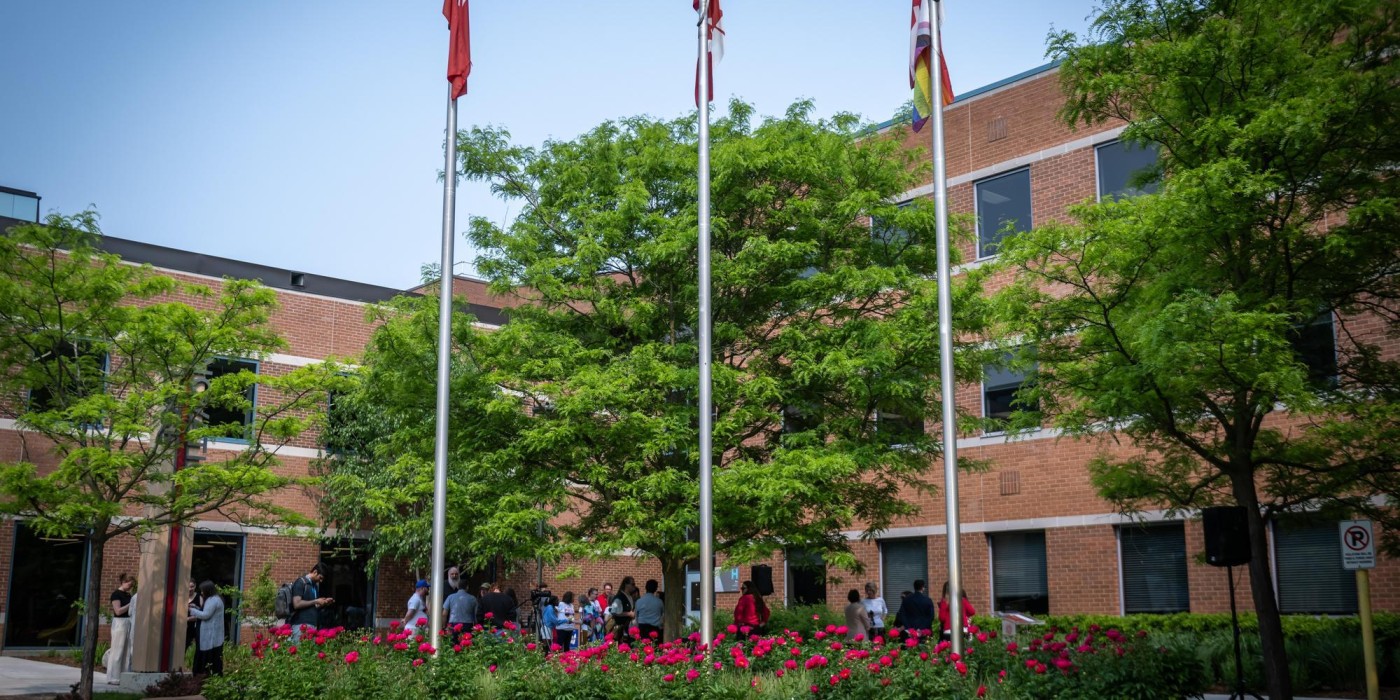 A red-brick building with large windows is surrounded by green trees and vibrant red flowers. Three flagpoles stand prominently, with a group of people gathered nearby.