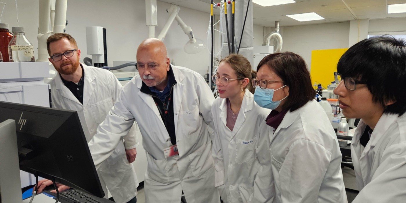 A group of five lab technicians in white coats gather around a computer in a bright laboratory. They appear focused and engaged, analyzing data on the screen.