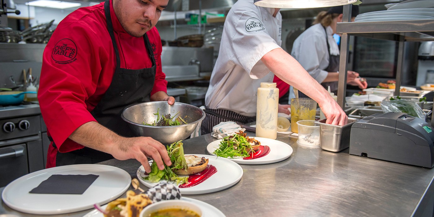 Two chefs in a commercial kitchen plate dishes, one in a red jacket arranging greens, the other in white preparing ingredients at a stainless steel counter.