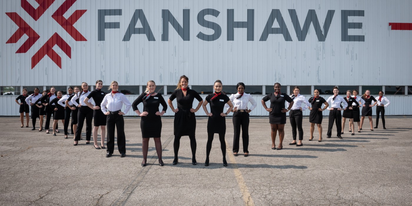 Faculty and students from the Flight Services program line up in uniform in front of the large sliding doors to the hangar which is decorated in a huge Fanshawe logo