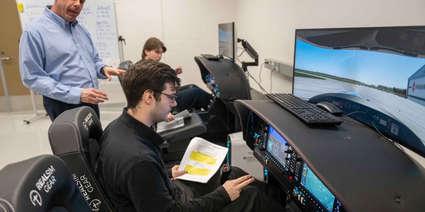 Inside a Fanshawe College classroom, individuals sit in flight simulator chairs with monitors displaying cockpit views, while an instructor stands nearby.