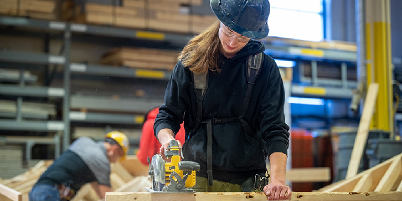 A female tradesperson cuts wood with a skill saw in a shop environment.