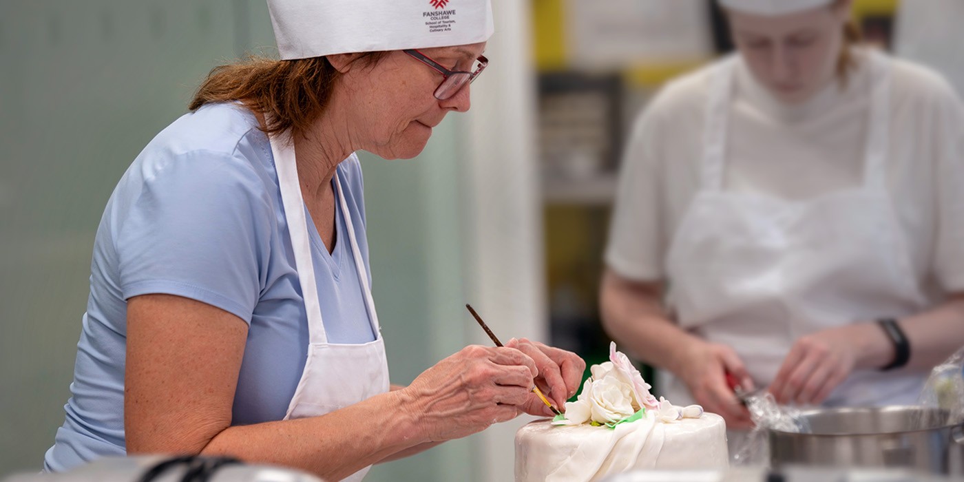 Two students in aprons carefully paint food colouring onto iced cakes