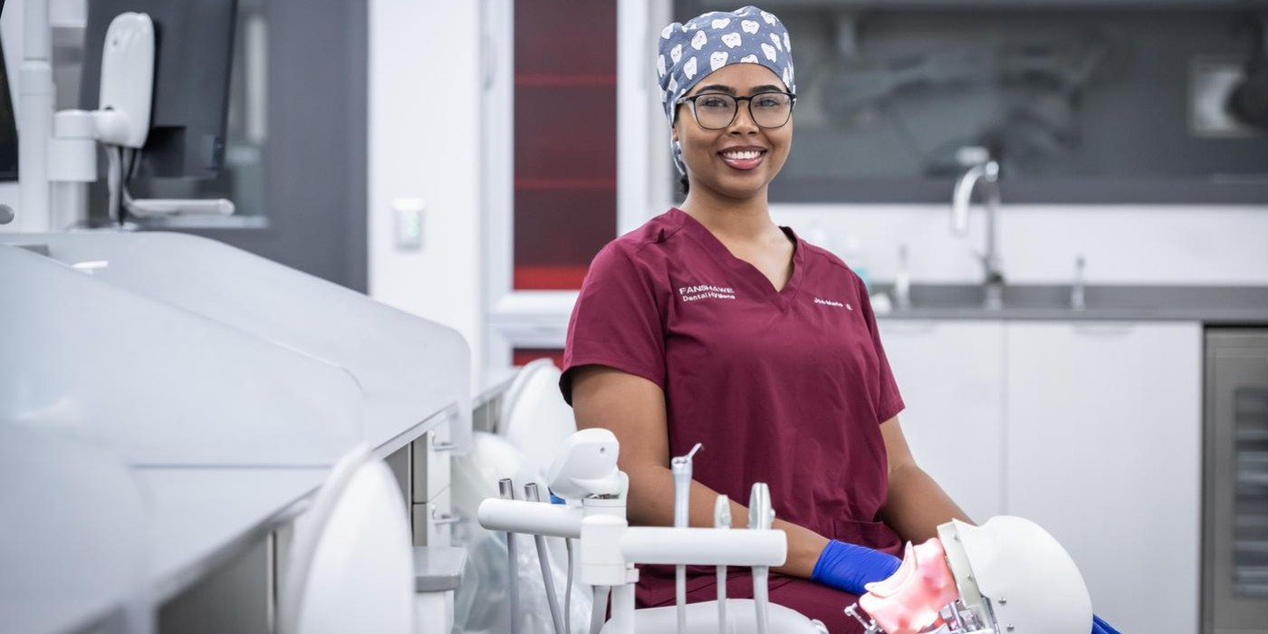 In the School of Health Sciences at Fanshawe College's London Campus, someone wearing a burgundy scrub top and a surgical cap patterned with teeth sits in a dental chair, with a dental mannequin in their lap.