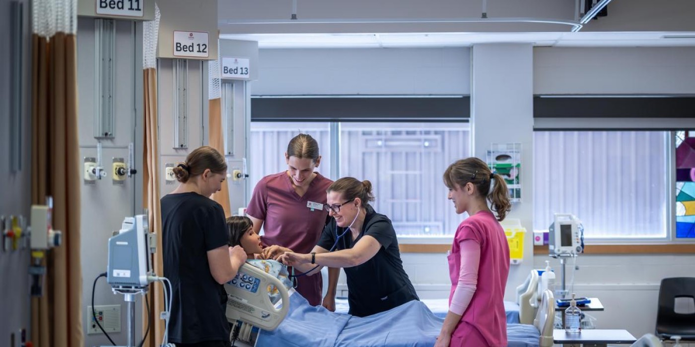 In a bright hospital room, four individuals in scrubs surround a patient in a bed with blue linens, one using a stethoscope