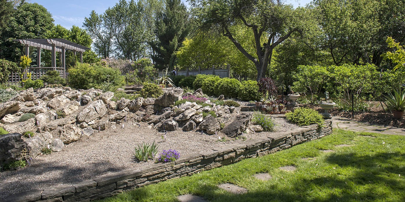 The Mac Cuddy Botanic Garden features a rock garden with a gravel bed, surrounded by a low stone wall and lush greenery, including a wooden pergola in the background.