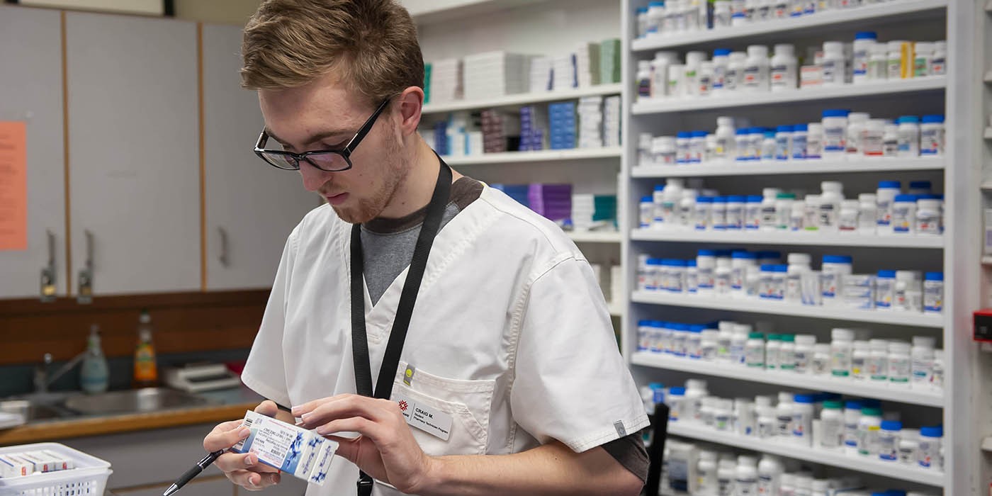 Inside the School of Health Sciences at Fanshawe College's London Campus, a person wearing glasses and a white lab coat holds a blister pack of medication and a pen, standing in front of shelves stocked with numerous bottles of pills.