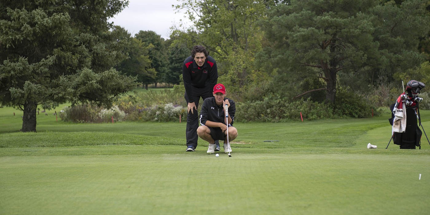 On a green golf course, one person crouches with a putter near a golf ball, while another person stands behind them, observing. A golf bag sits nearby, and trees line the background.