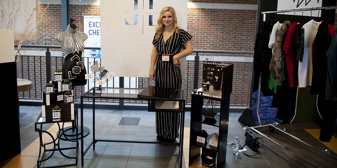 At the London Campus of Fanshawe College, a person stands in front of a display for the Fashion Marketing and Management program, featuring a banner with a logo, a mannequin, miniature displays, and a clothing rack.