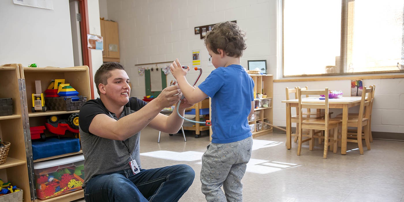 At the Huron-Bruce Regional Site of Fanshawe College in Clinton, someone sits on a rug in the Early Childhood Education program classroom, holding a toy stethoscope to a child's chest. A toy tool box sits on the rug, and shelves of toys are in the background. A small table and chairs sit near a window.