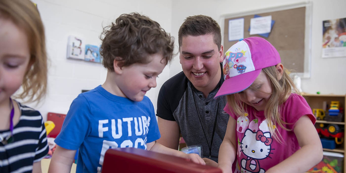 Inside a classroom at the Huron-Bruce Regional Site of Fanshawe College in Clinton, an Early Childhood Education program event shows a smiling person with short brown hair, wearing a lanyard, leaning over a red toy with two children. One child wears a blue shirt with the word "FUTURE" on it, and the other child wears a pink shirt and a pink baseball cap. A clock hangs on the wall in the background.