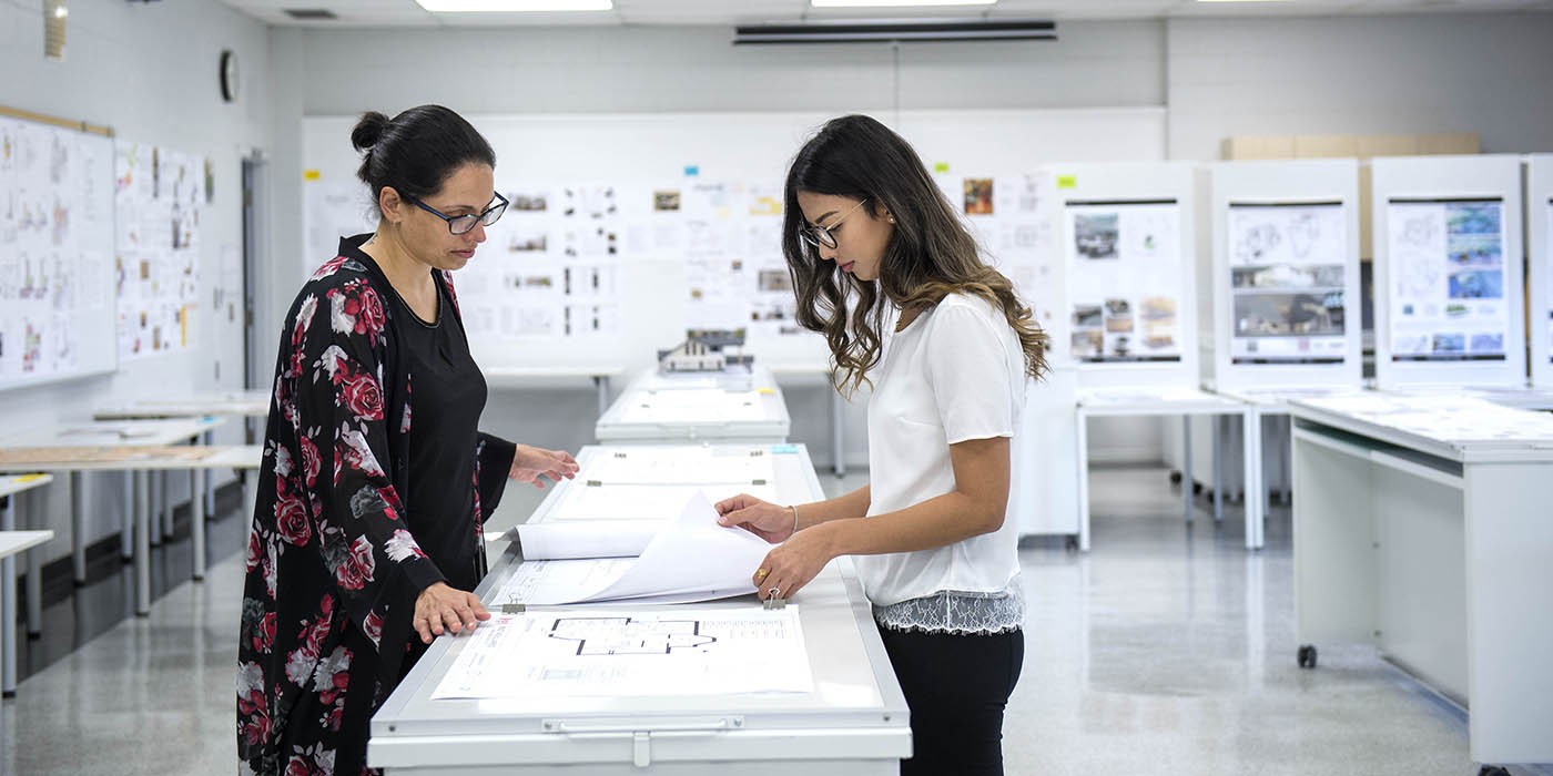 Two people stand at a table reviewing architectural drawings in a bright classroom at the London Campus of Fanshawe College. Whiteboards and presentation boards line the walls.