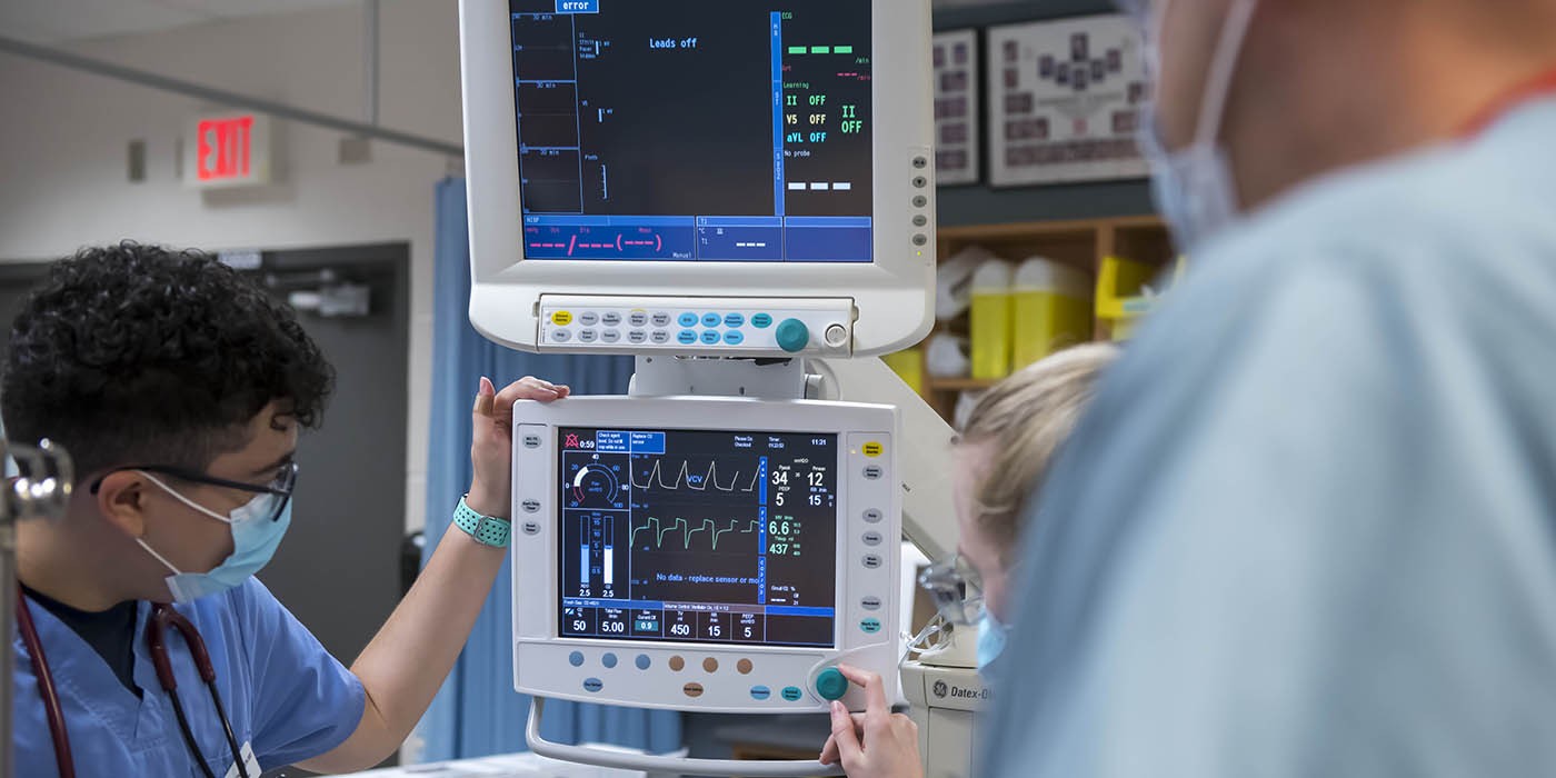 At the School of Health Sciences of Fanshawe College at the London Campus, two people in scrubs and masks are operating a medical ventilator, which has two screens displaying various readings and settings.
