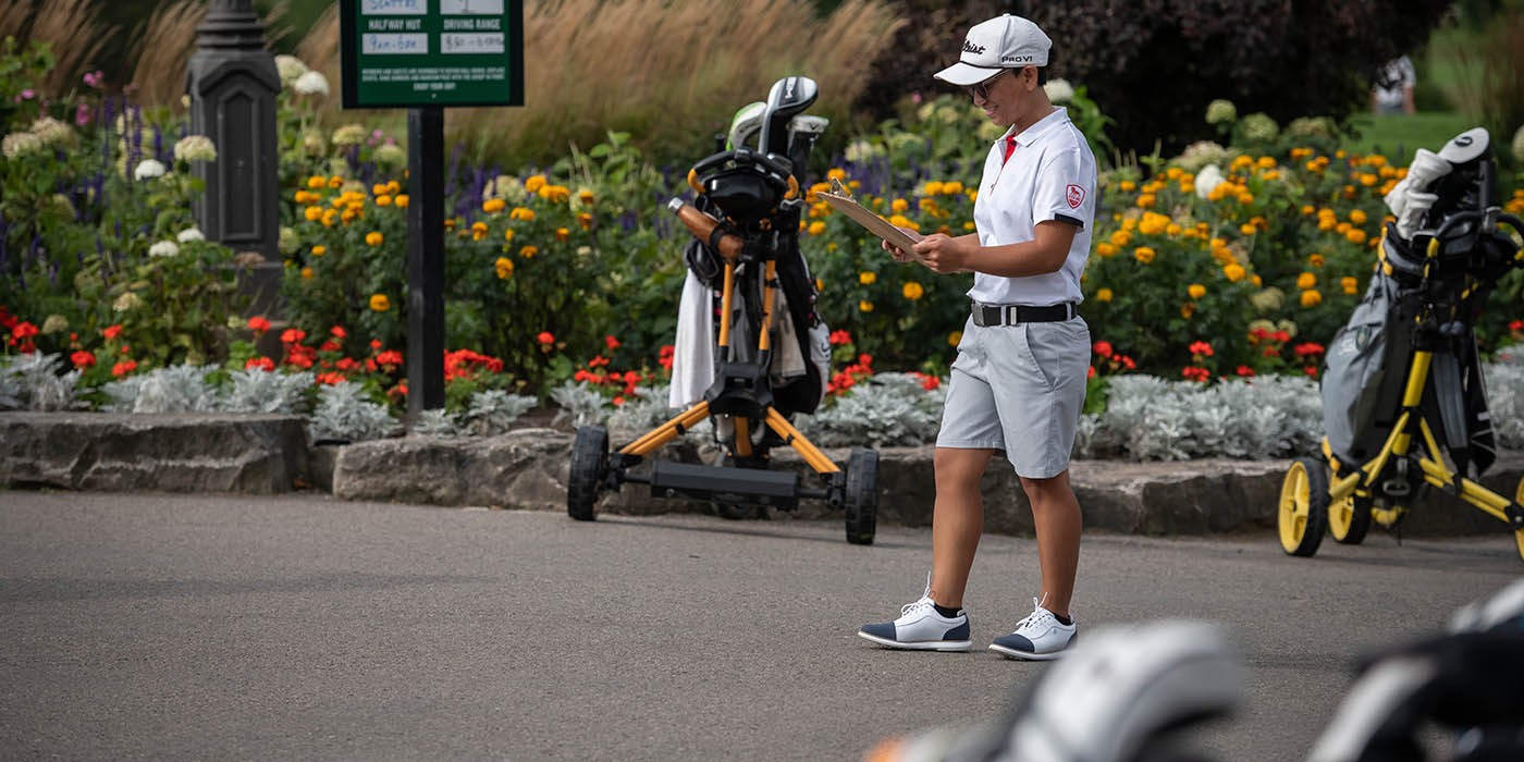 At a golf course, a person in a white polo shirt, grey shorts, and a white cap stands on a paved path, holding a clipboard, with golf bags and carts nearby, and a course status sign visible behind a flower bed.