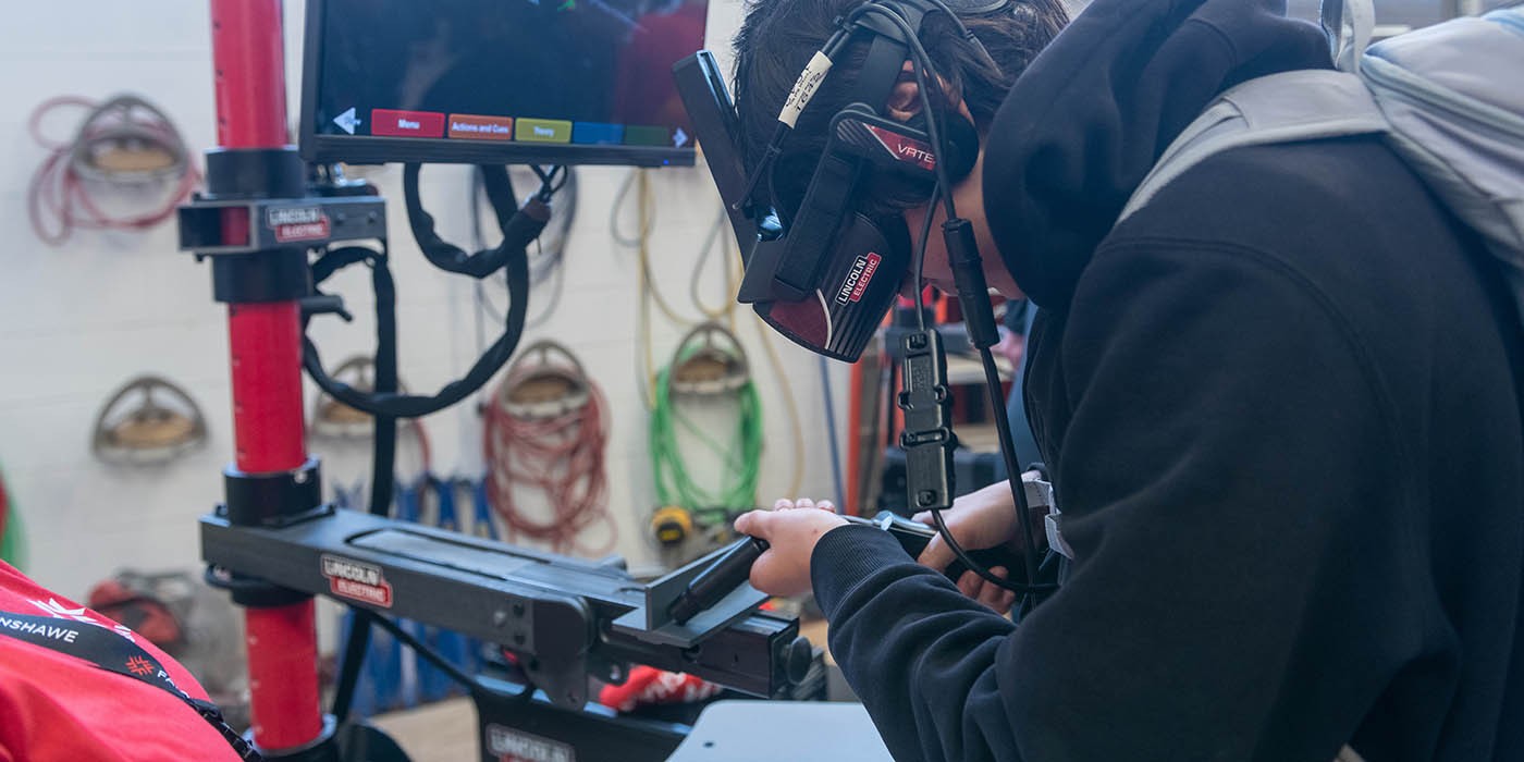 A person uses a virtual reality welding simulator at Fanshawe College, with a Lincoln Electric monitor displaying a welding simulation.