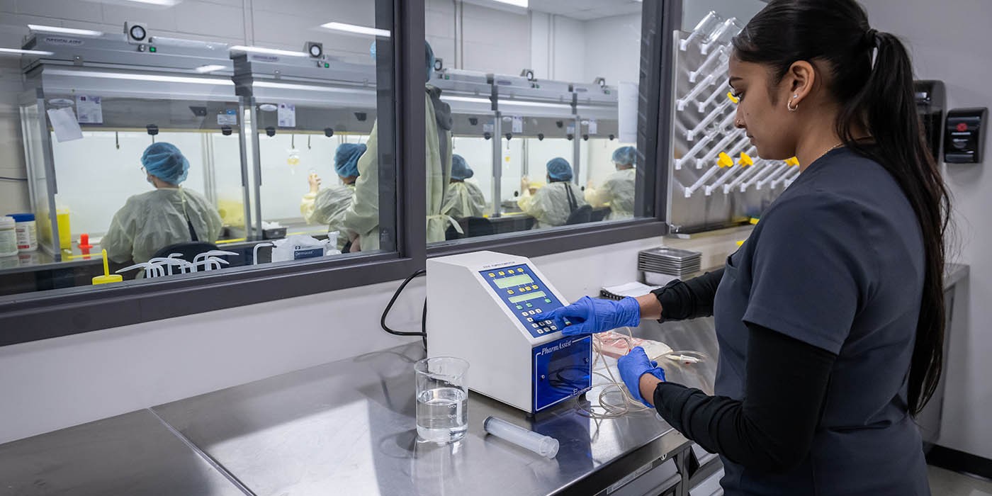 At the School of Health Sciences of Fanshawe College at the London Campus, a person wearing a grey uniform with a Fanshawe College Community Pharmacy Assistant Student patch is pressing a button on a keypad.
