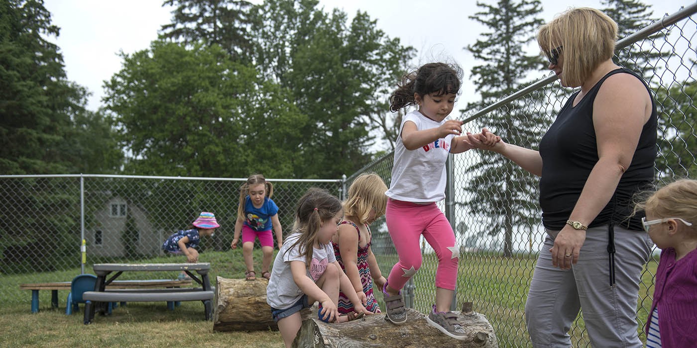 Early Childhood Education program participants are seen on the Simcoe/Norfolk Regional Campus, engaging in outdoor play on a log obstacle course, with a chain link fence and trees in the background.