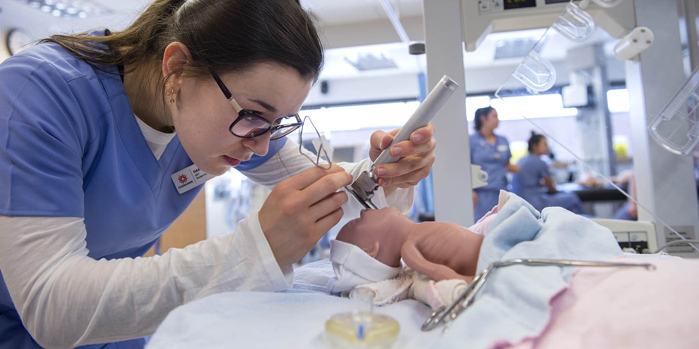 In the School of Health Sciences at Fanshawe College's London Campus, a person wearing a blue scrub top and glasses uses medical instruments to examine a baby mannequin lying on a bed with blue and pink blankets.