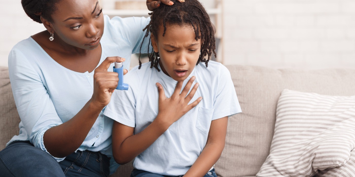 a child sits on a couch struggling to breathe next to his mother who is holding his asthma inhaler prepared to support him.