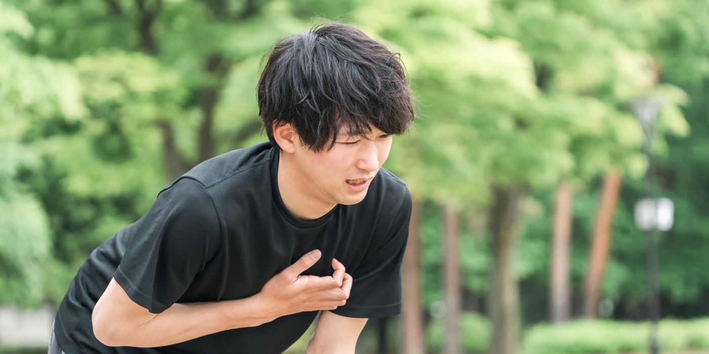 A young male experiences breathing difficulty while on a run outdoors. He appears bent over with a hand on his chest attempting to catch his breathe with blurred trees behind him.