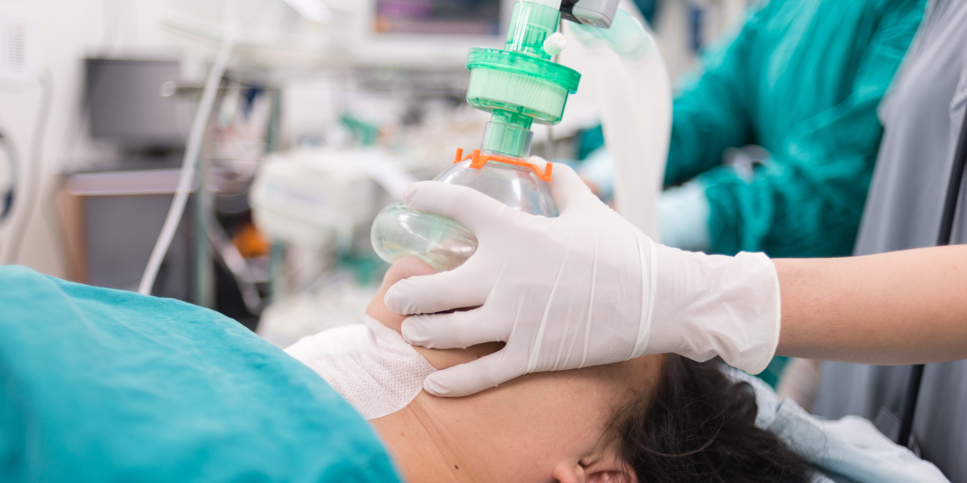A patent on an operating room table, with a breathing mask over their face being hold in place by an RT