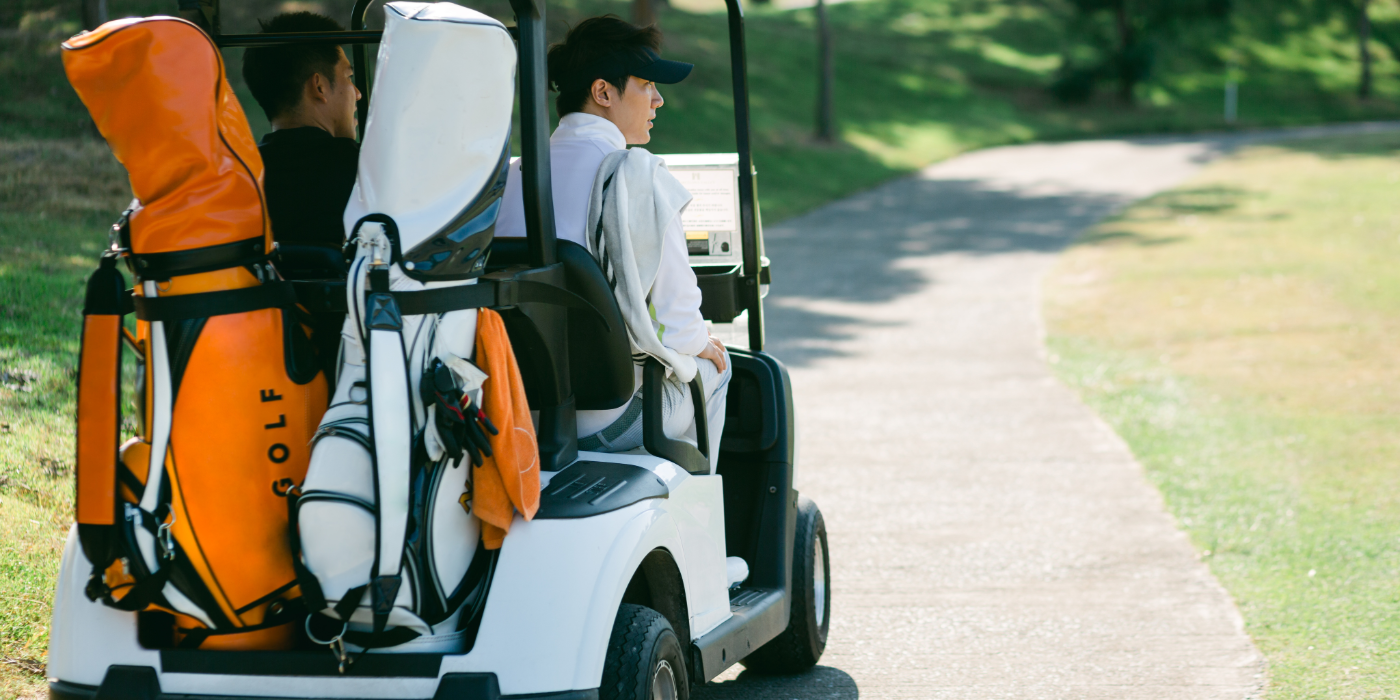 A golf cart on a course pathway, with two golf bags on the back, one orange and one white, two golfers sit in the front